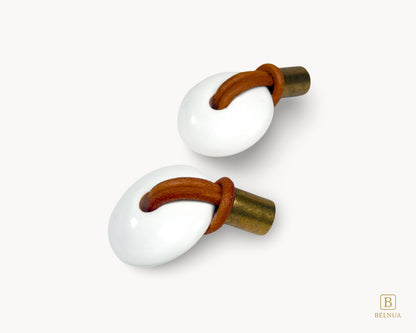 A pair of white porcelain and antique brass drawer knobs featuring rustic leather loops for a vintage aesthetic.