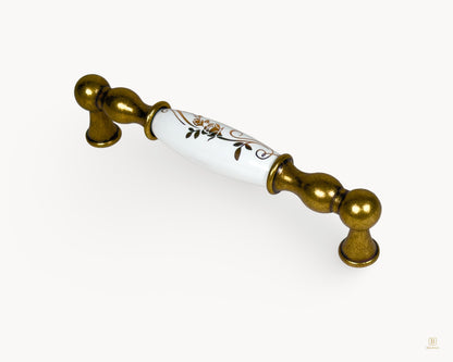 Angled view of Belmira antique brass handle with white porcelain and golden scroll detailing, 128mm