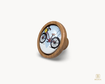 Belnua - Front left angled view of delicate Nuvino children knob with natural oak wood base and bicycle design porcelain insert for boys, girls, nursery, school, teens room and furniture