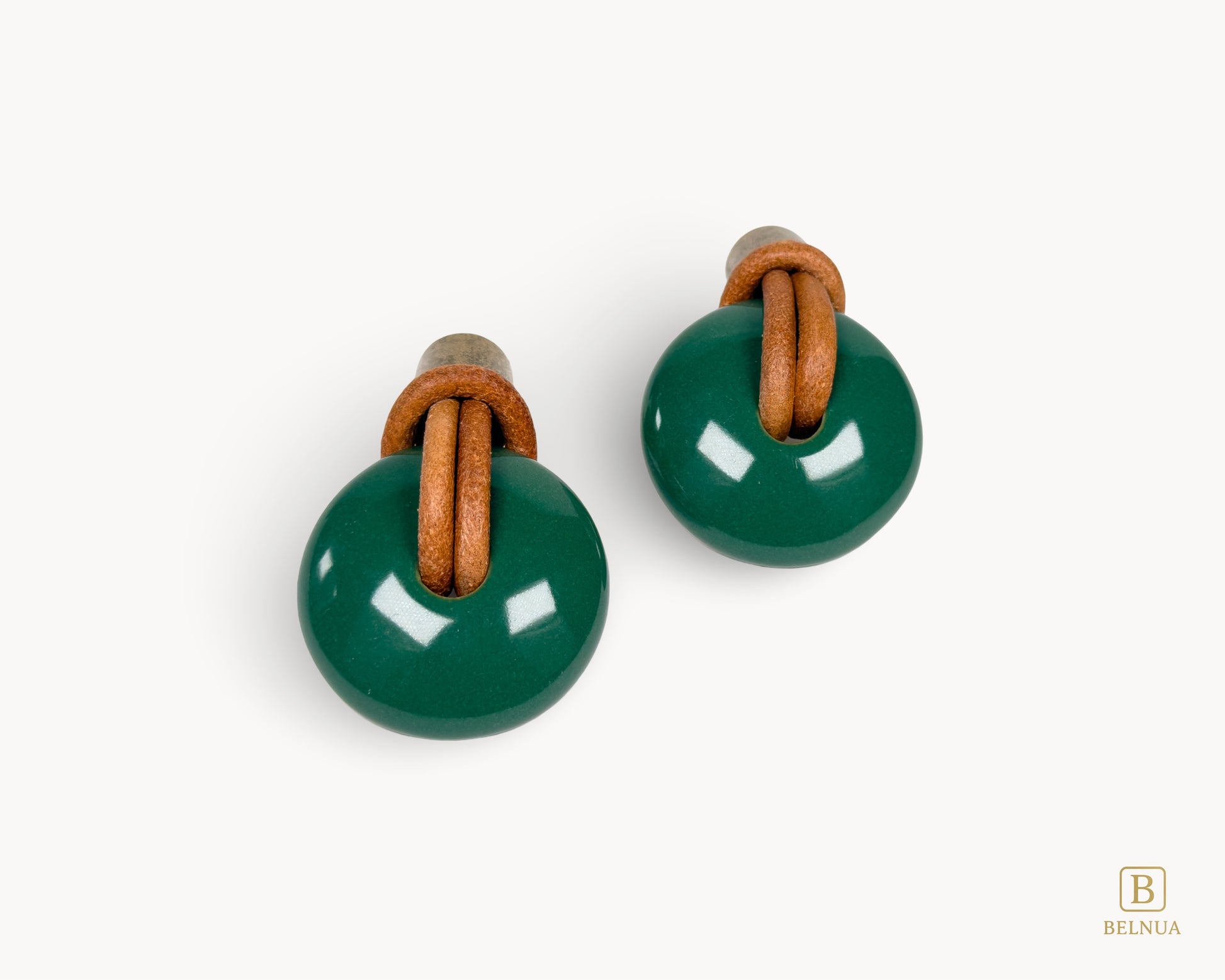 Front view of a pair of Belnua Revina dark green porcelain knobs with antique brass hardware and leather loops.