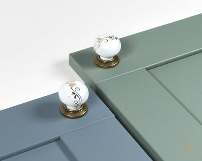 Belnua - ZERRA porcelain/ceramic white knob with golden scroll motif with antique brass base on dusty blue and sage green cabinet door